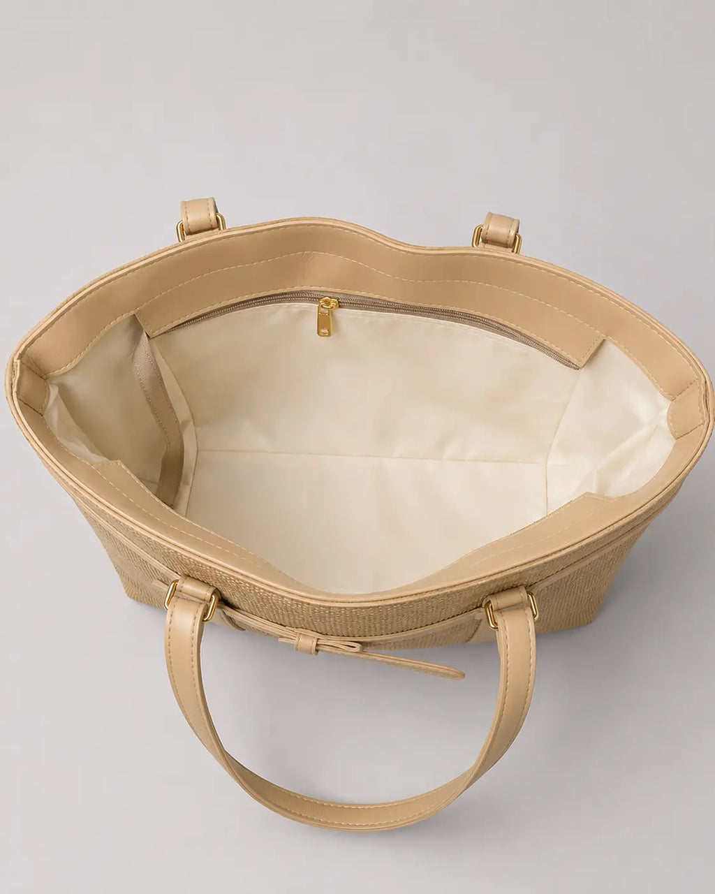 A structured tote bag in a light beige textured leather, shown from an overhead angle to display its cream-colored interior lining and double top handles against a neutral gray background.