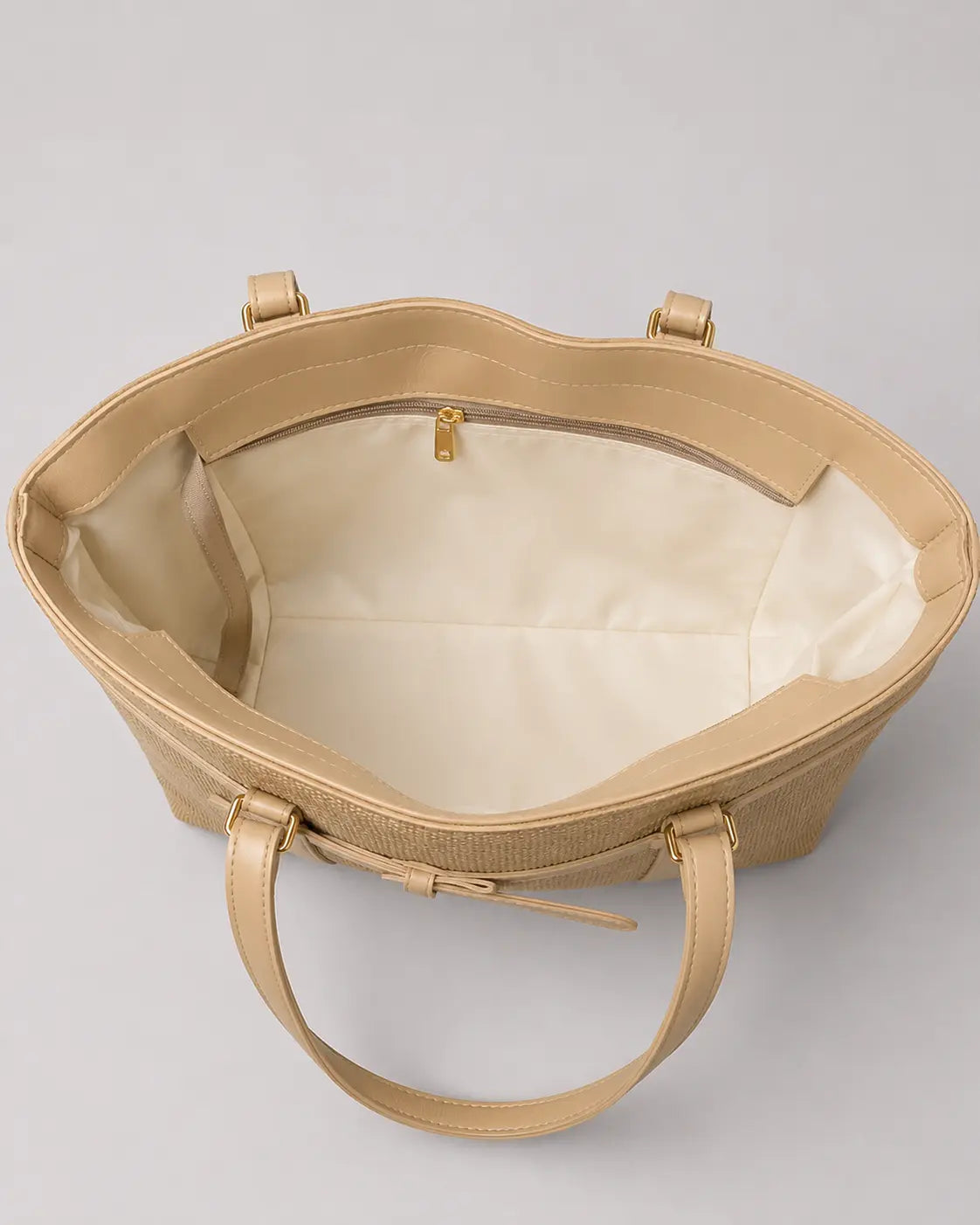 A structured tote bag in a light beige textured leather, shown from an overhead angle to display its cream-colored interior lining and double top handles against a neutral gray background.