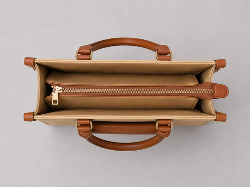 Top-down view of a structured tote bag with tan canvas exterior, warm brown leather handles, and a gold-tone zipper closure, shown on a neutral gray background.