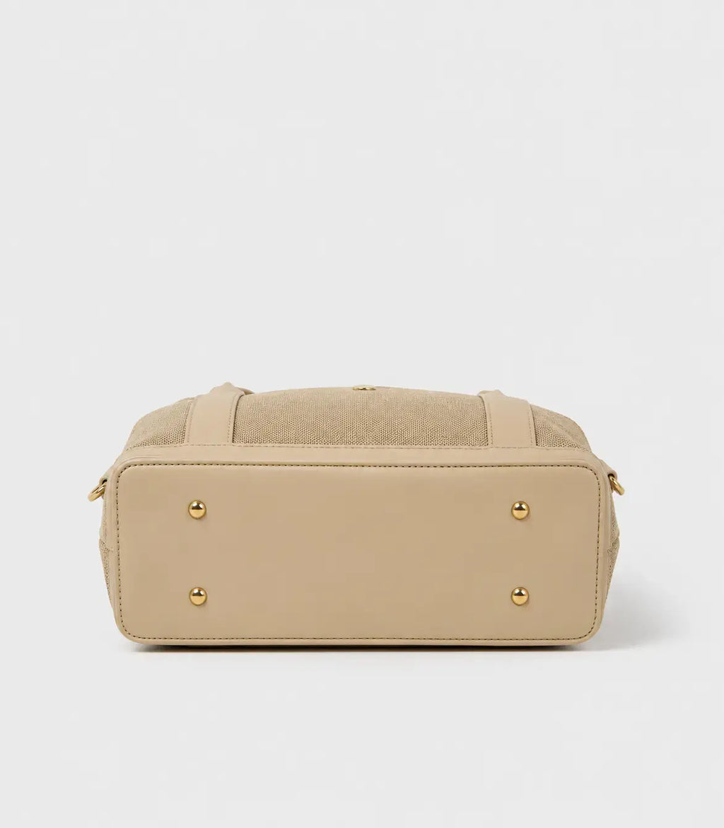 A beige handbag shown from the back, featuring a smooth leather base, a textured fabric top panel, and four gold-tone metal studs on the back corners.