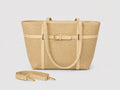 A structured tote bag in a woven beige material with light beige trim, featuring a central buckle strap detail and a detachable shoulder strap placed in front, set against a plain light gray background.