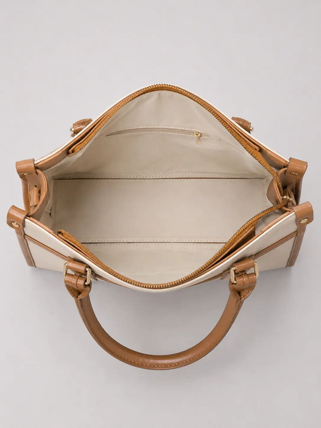 An open top view of a structured handbag with cream-colored panels and tan leather trim, handles, and zippers, set against a plain light gray background.