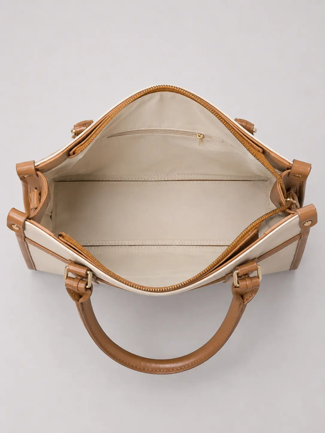 An open top view of a structured handbag with cream-colored panels and tan leather trim, handles, and zippers, set against a plain light gray background.