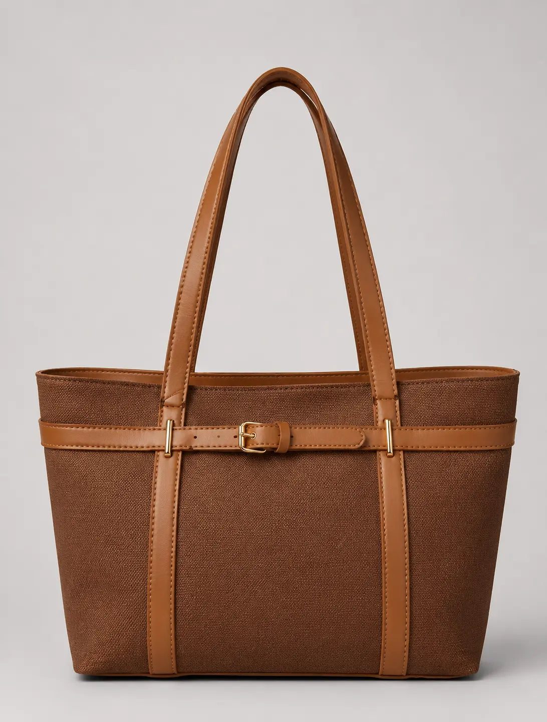 A structured brown tote bag with tan leather handles, trim, and a central buckle strap detail, photographed against a neutral light gray background.
