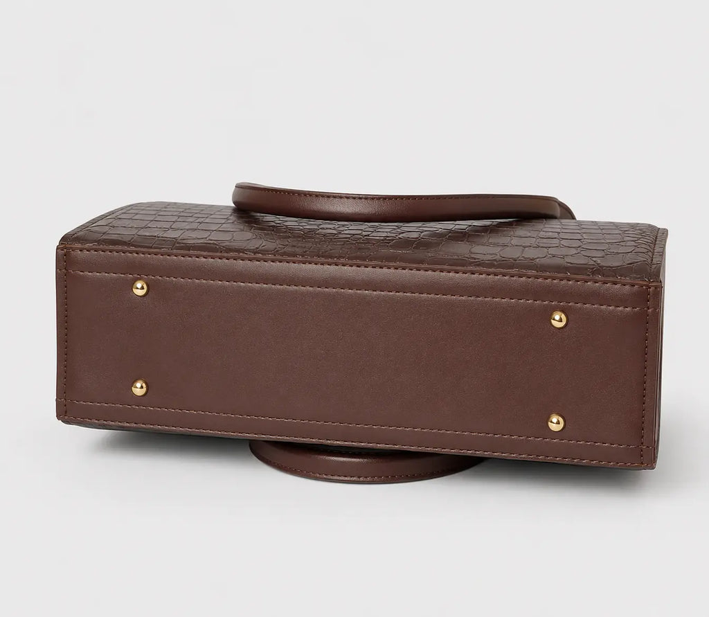 A brown rectangular handbag with a crocodile-embossed top panel, smooth brown leather handle and trim, and small gold-tone metal studs on the front, shown on a light gray background.