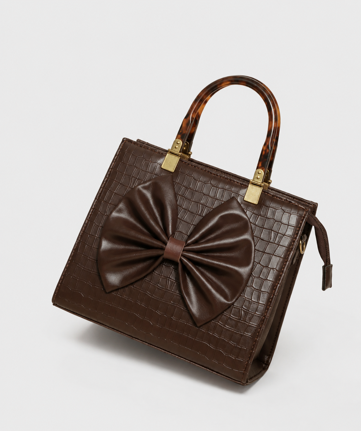 A dark brown crocodile-embossed leather handbag with a large matching bow on the front, dual top handles in a tortoiseshell pattern, and gold-tone hardware, shown on a light gray background.