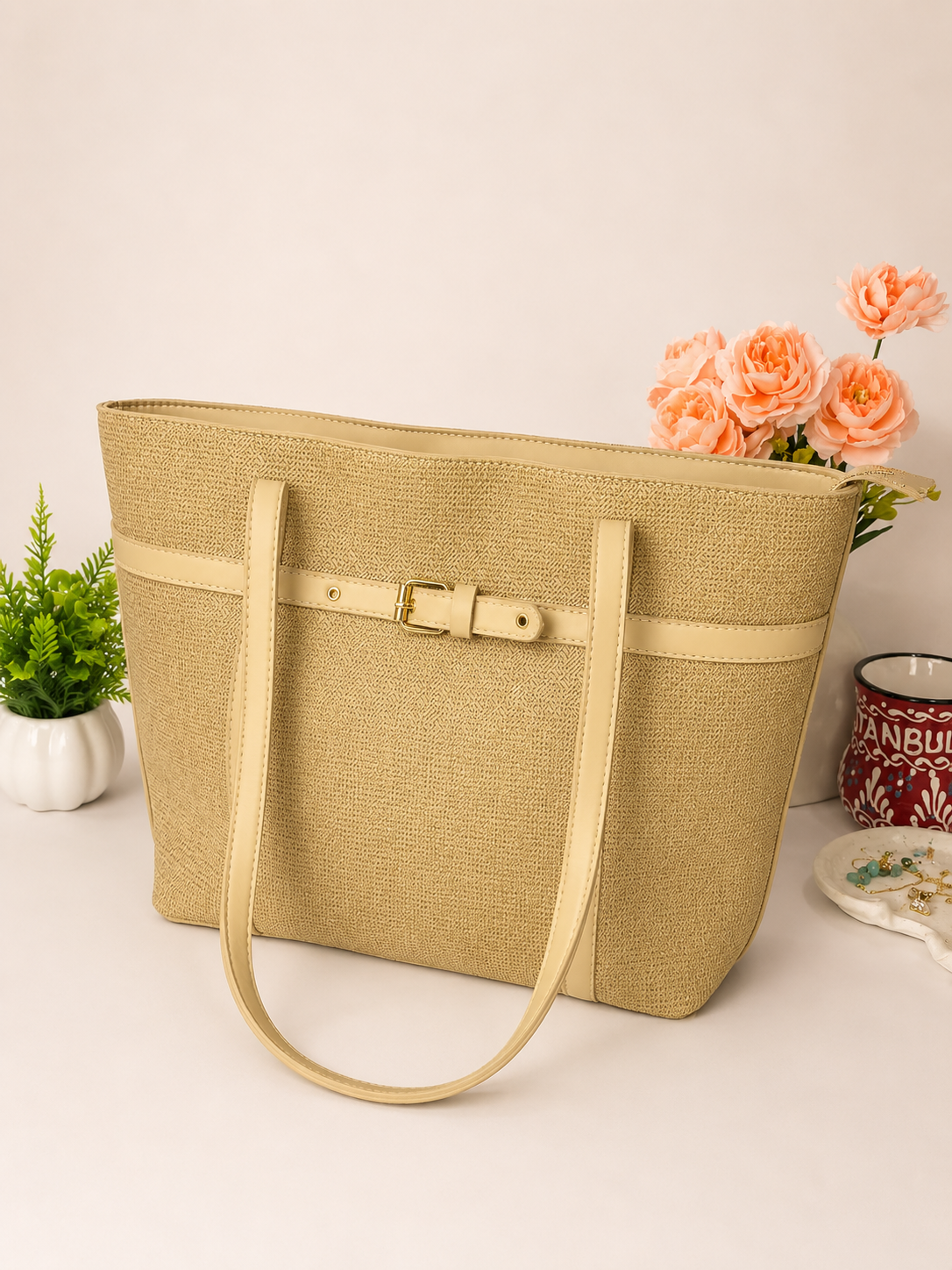 A beige woven tote bag with a matching leather strap and buckle detail, styled on a light surface with decorative plants and flowers in the background.