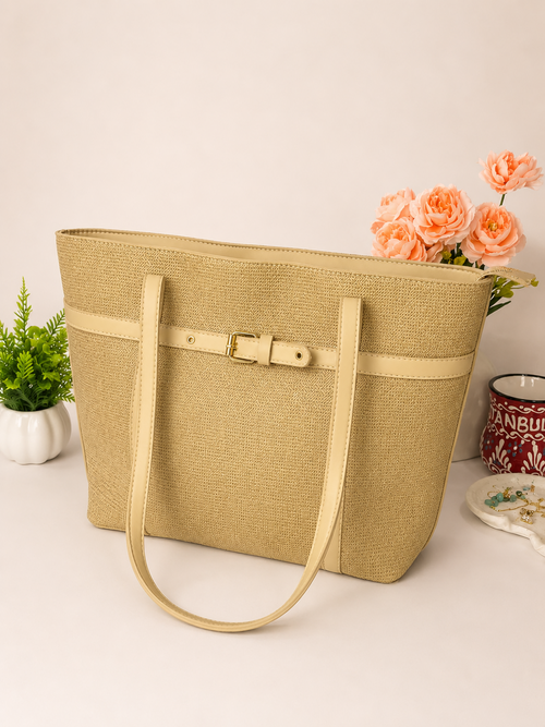 A beige woven tote bag with a matching leather strap and buckle detail, styled on a light surface with decorative plants and flowers in the background.