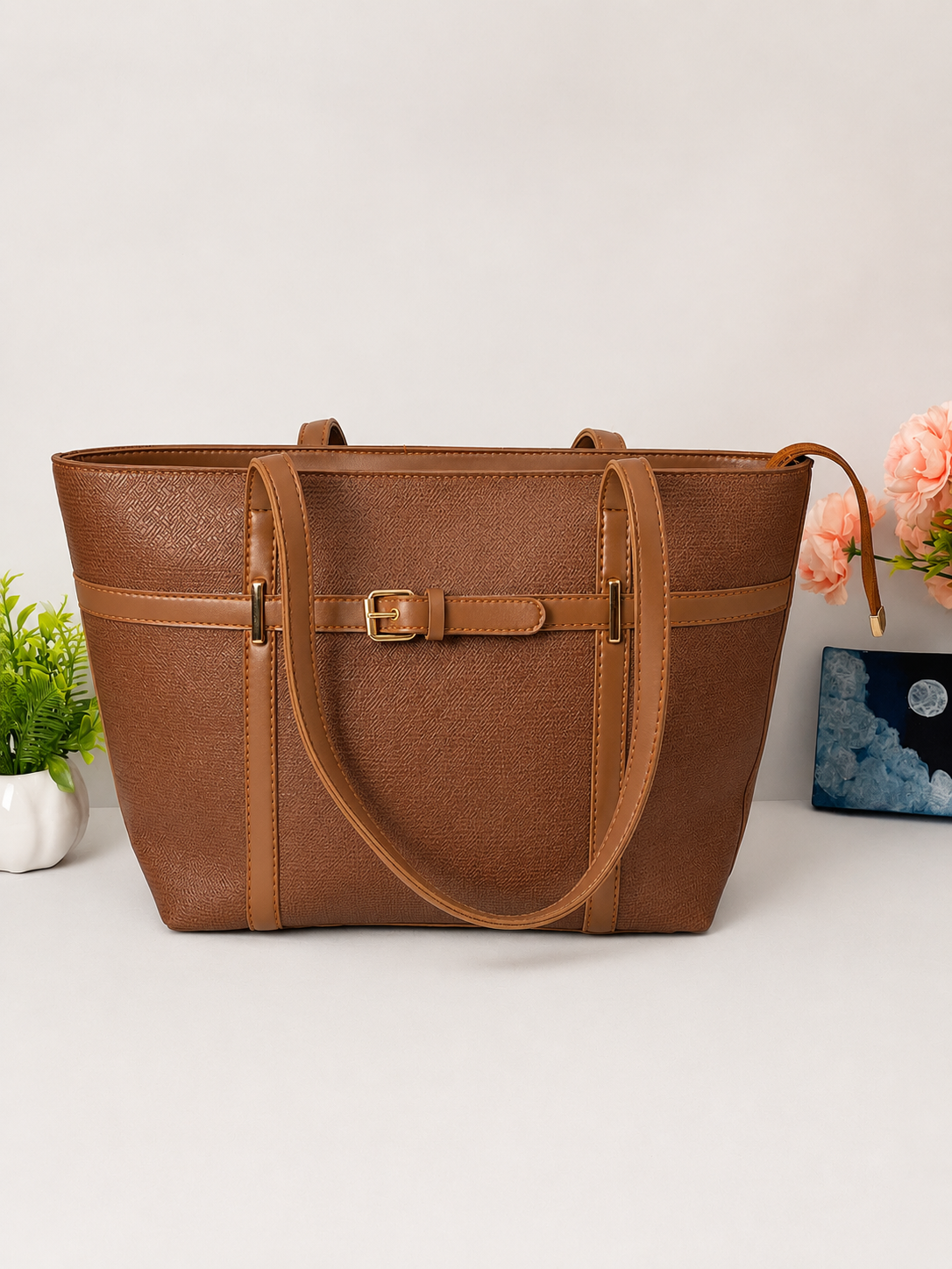 A structured brown faux-leather tote bag with a central horizontal strap and buckle detail, displayed on a light gray surface with small decorative plants and flowers.