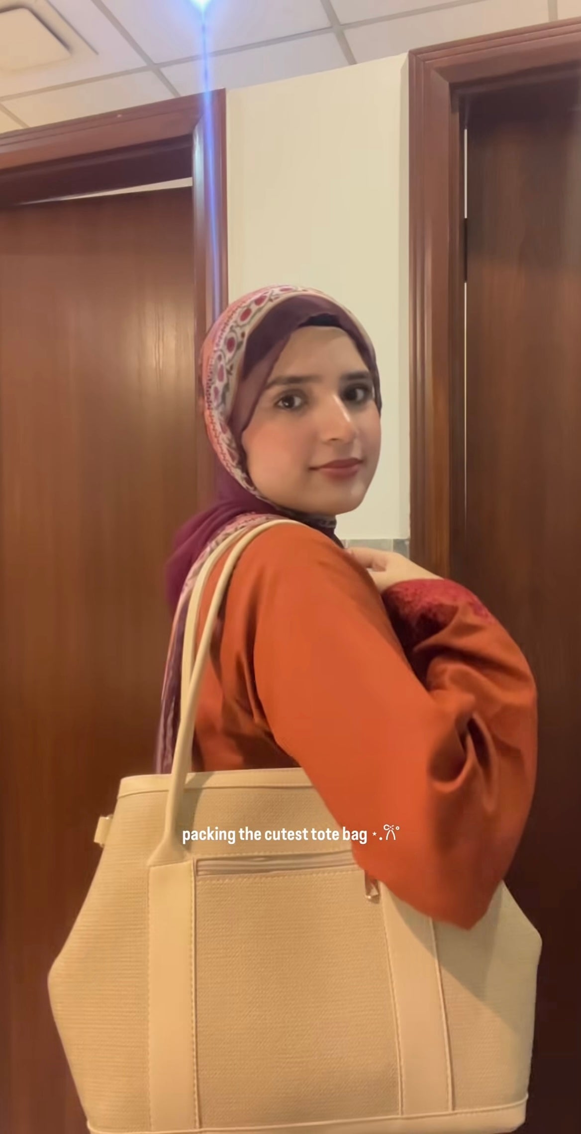 A woman wearing a patterned hijab and an orange-brown top carries a large cream-colored tote bag over her shoulder in an indoor setting with wooden doors.