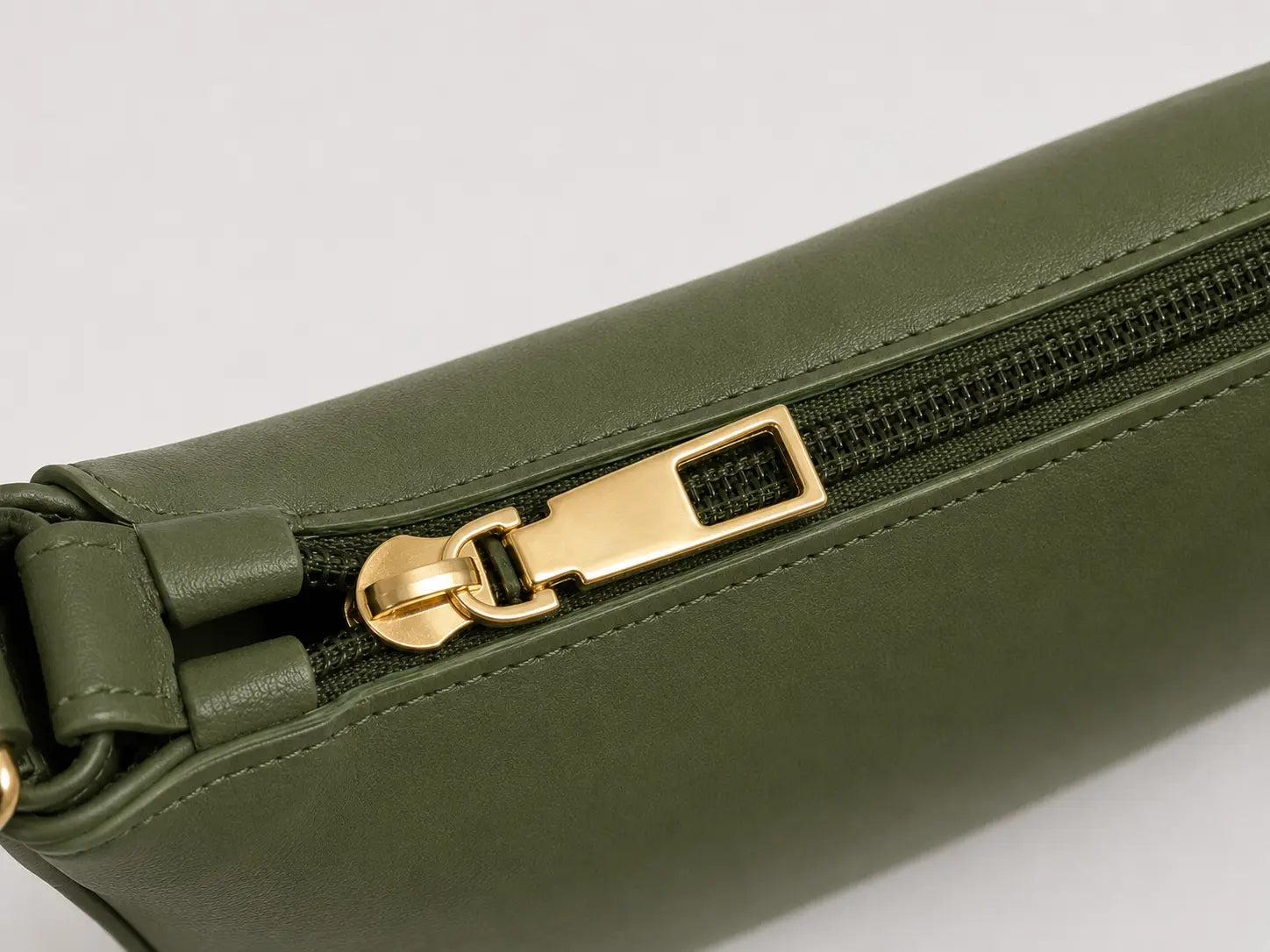 Close-up of an olive green leather zippered pouch with a gold-tone metal zipper pull on a light gray background.