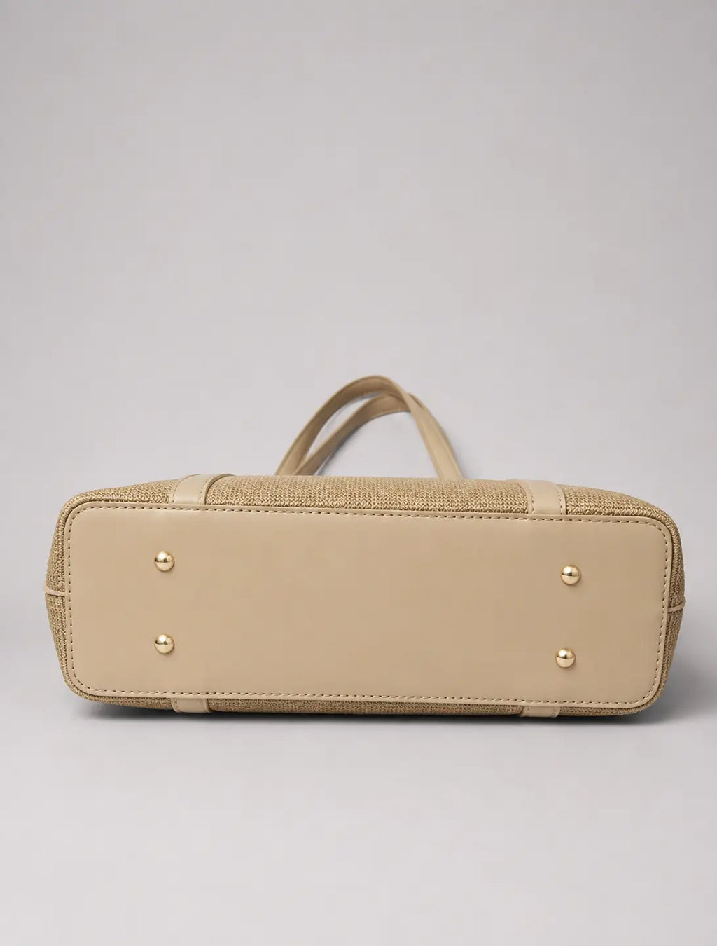 A beige leather and woven jute handbag shown from the bottom, featuring a smooth leather base, textured woven side panels, top handles, and gold-tone metal studs, set against a neutral gray background.