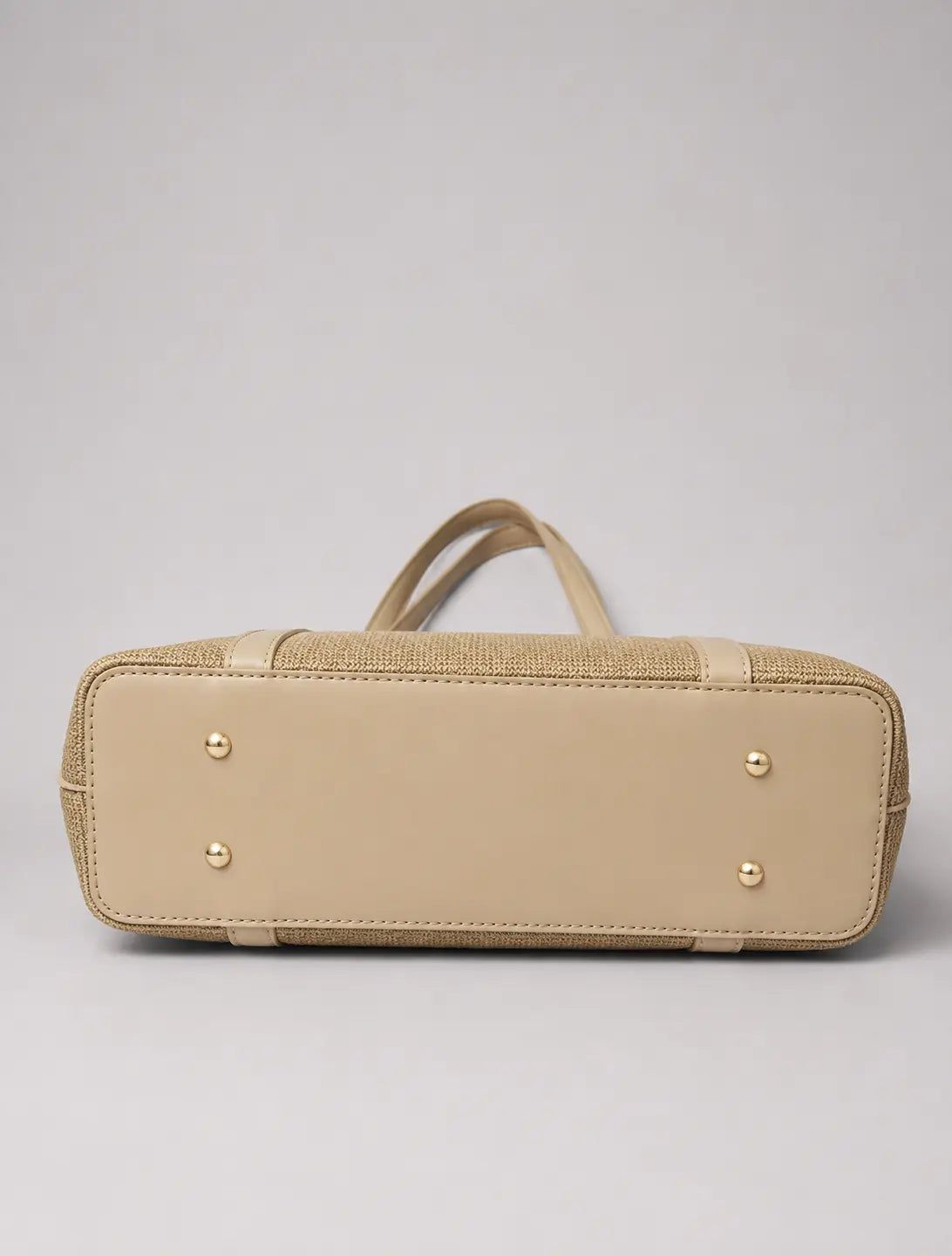 A beige leather and woven jute handbag shown from the bottom, featuring a smooth leather base, textured woven side panels, top handles, and gold-tone metal studs, set against a neutral gray background.