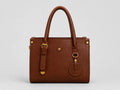 A structured brown leather handbag with dual top handles, gold-tone hardware, and a circular leather charm, photographed against a neutral light gray background.