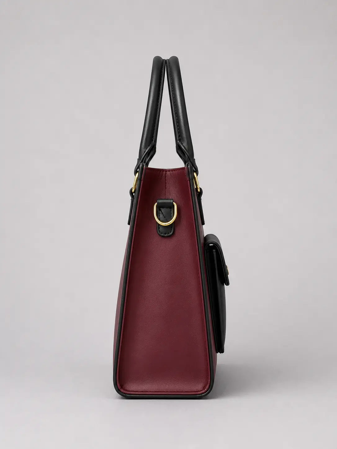A structured tote bag in burgundy leather with black leather handles and trim, featuring gold-tone hardware, shown from a side profile view against a neutral gray background.
