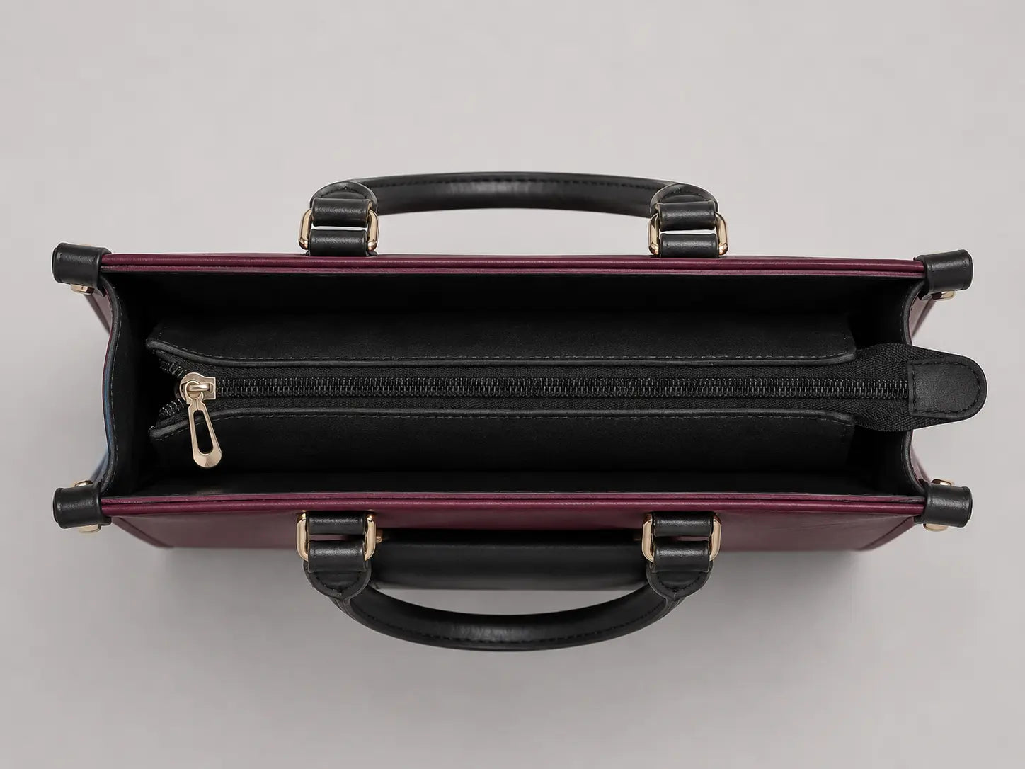 Top-down view of a structured handbag in burgundy and black leather, showing the interior zippered compartment and gold-tone hardware against a light gray background.
