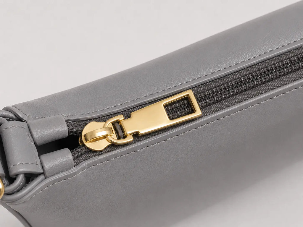 Close-up of a gray leather zipper pouch with a gold-tone metal zipper pull on a light gray background.