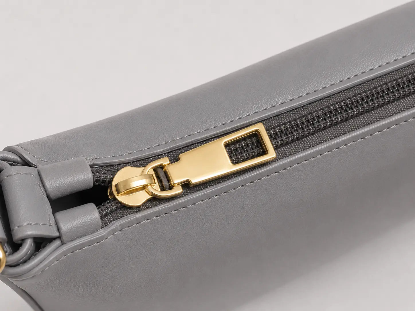 Close-up of a gray leather zipper pouch with a gold-tone metal zipper pull on a light gray background.