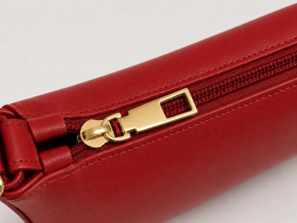 A close-up of a red leather zippered pouch or clutch with a polished gold-tone metal zipper pull on a light gray background.