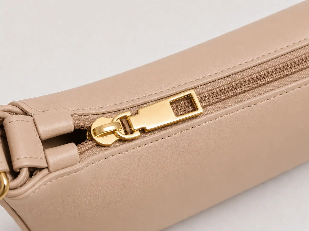 Close-up of a beige leather zippered pouch with a gold-tone metal zipper pull on a light gray background.