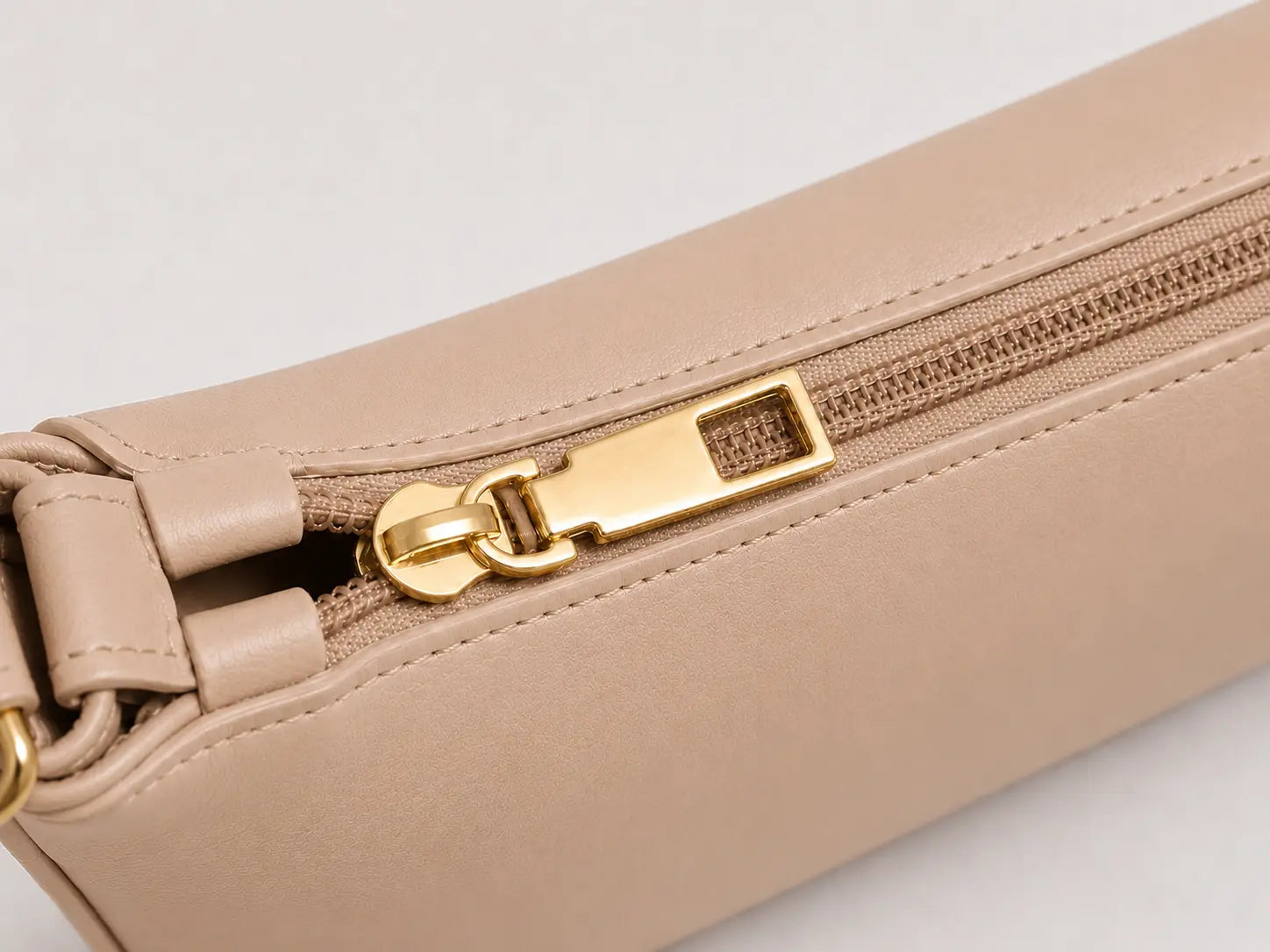 Close-up of a beige leather zippered pouch with a gold-tone metal zipper pull on a light gray background.