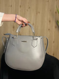 A light gray leather handbag with top handles and gold-tone hardware, held by a person's hand against a light wood panel background.