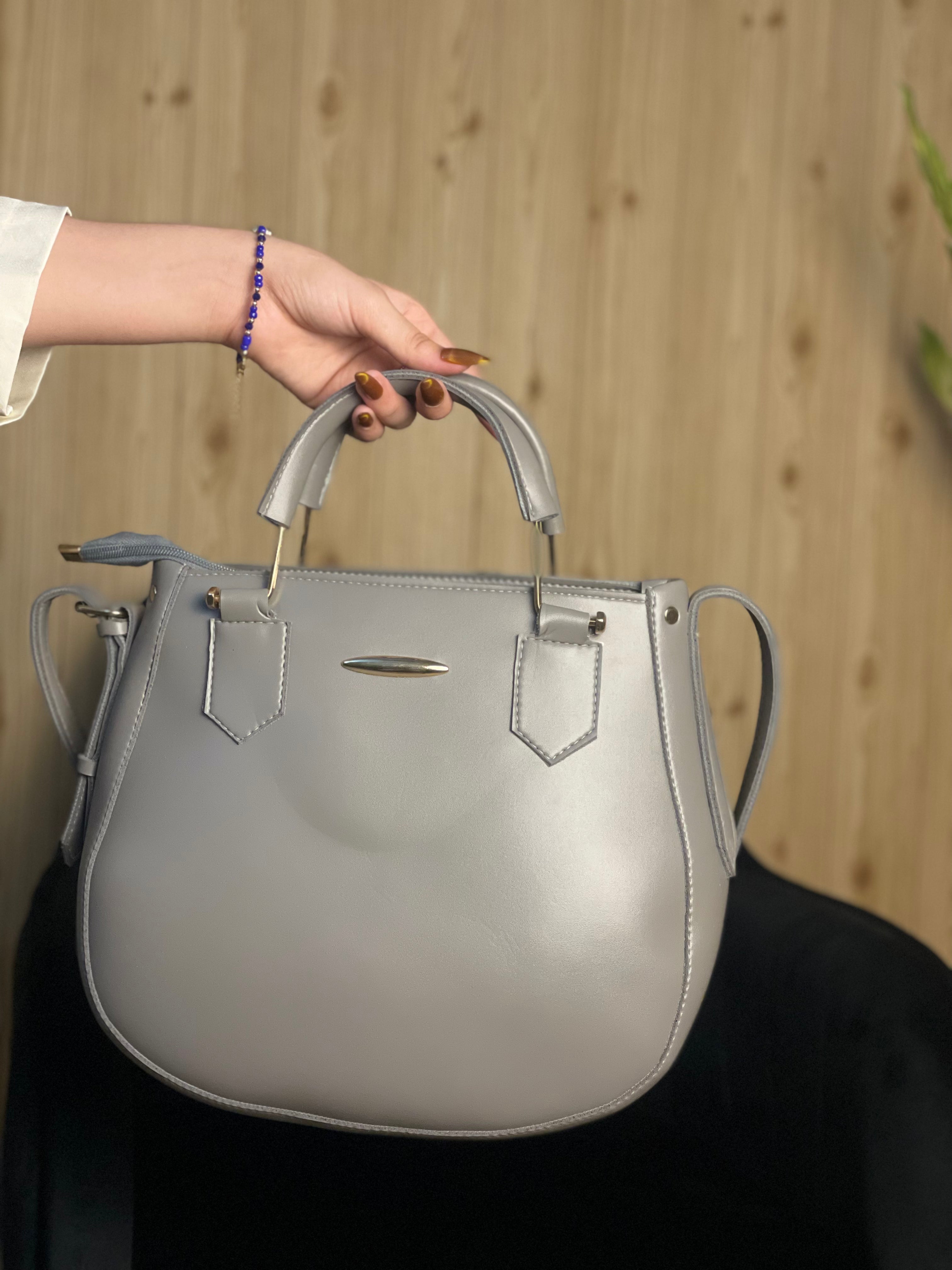 A light gray leather handbag with top handles and gold-tone hardware, held by a person's hand against a light wood panel background.